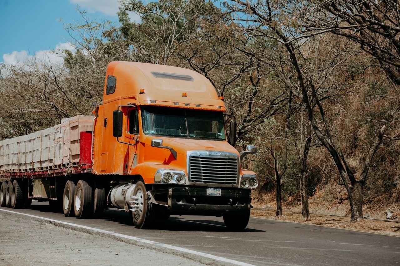 Free A vibrant orange Freightliner truck drives on a highway through a dry, scenic landscape in El Salvador. Stock Photo