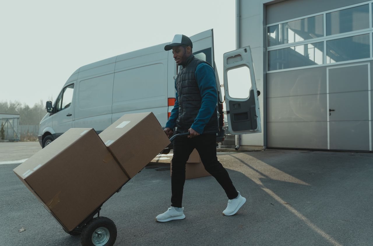 Free Courier pushing a trolley with boxes near a van at a warehouse, showcasing delivery services. Stock Photo
